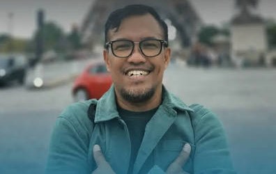 Friendly portrait of a man with glasses wearing a teal jacket, arms crossed, outdoors with a blurred city street behind him.