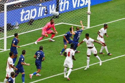 Soccer scene at the goal: players in blue and white contest the ball near the net as the pink-clad goalkeeper guards the goal.