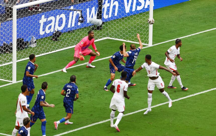 Soccer scene at the goal: players in blue and white contest the ball near the net as the pink-clad goalkeeper guards the goal.
