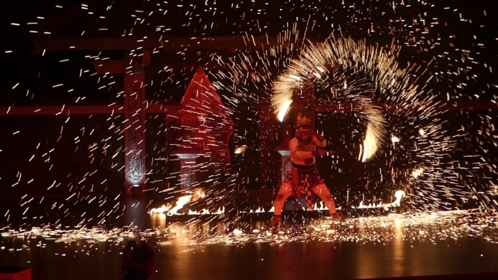 Fire dancer in red costume swinging flaming torches, sparks flying in a dark stage setting.