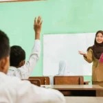 Teacher in a hijab speaks at the front of a classroom, holding a pink book while students raise hands to answer.