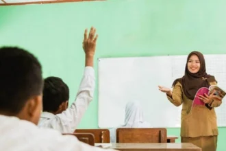Teacher in a hijab speaks at the front of a classroom, holding a pink book while students raise hands to answer.