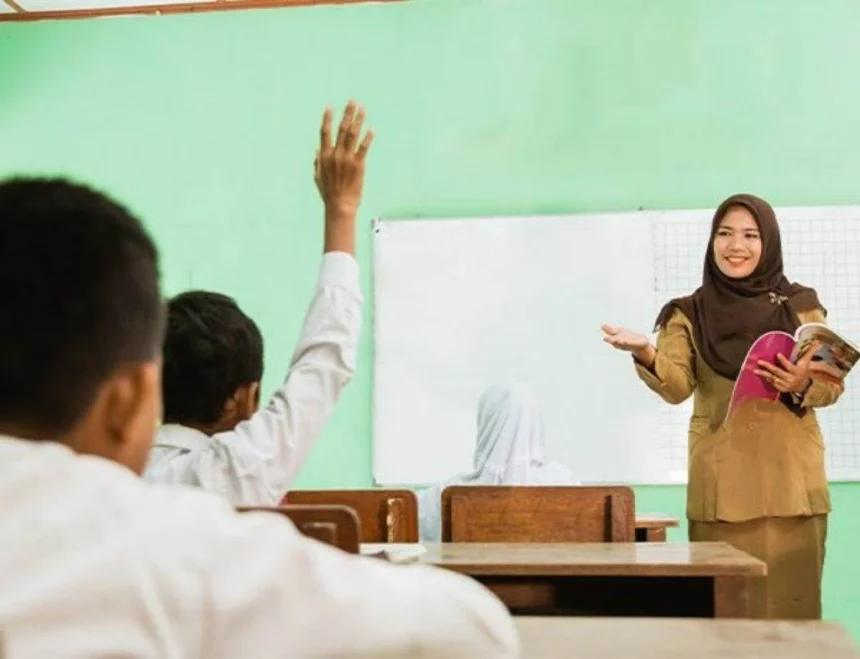 Teacher in a hijab speaks at the front of a classroom, holding a pink book while students raise hands to answer.