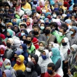 Large diverse crowd wearing face masks outdoors, standing close together.