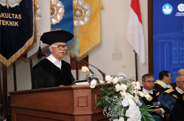 faAS - Mabur.co Professor in graduation gown and cap giving a speech at a wooden podium adorned with white flowers