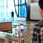 Traveler with a backpack stands at a metal security gate with a smartphone facial-recognition kiosk mounted on a stand in a modern terminal, foregrounded by another traveler.