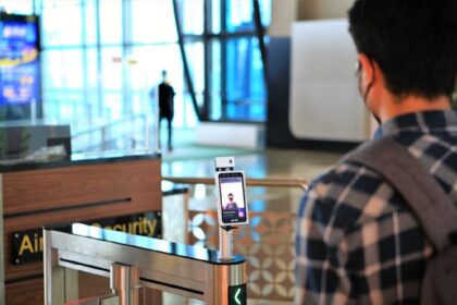 Traveler with a backpack stands at a metal security gate with a smartphone facial-recognition kiosk mounted on a stand in a modern terminal, foregrounded by another traveler.