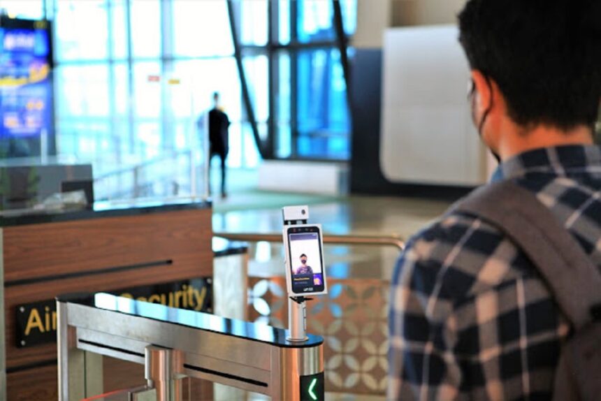 Traveler with a backpack stands at a metal security gate with a smartphone facial-recognition kiosk mounted on a stand in a modern terminal, foregrounded by another traveler.