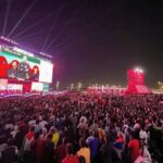 Massive outdoor concert crowd at night, watching a colorful stage with giant video screens.