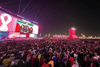 Massive outdoor concert crowd at night, watching a colorful stage with giant video screens.