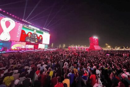 Massive outdoor concert crowd at night, watching a colorful stage with giant video screens.