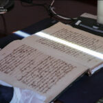 Open antique manuscript with dense handwritten text on a desk; gloved hands and a scanning device nearby.