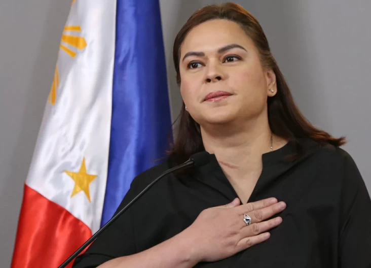 Woman speaking at a podium with her hand over her chest, the Philippine flag in the background with soft lighting.