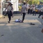 Police officers in tactical gear on a street, debris scattered on the pavement, a crumpled cloth on the ground, and an officer in the foreground handling a rope or cord.