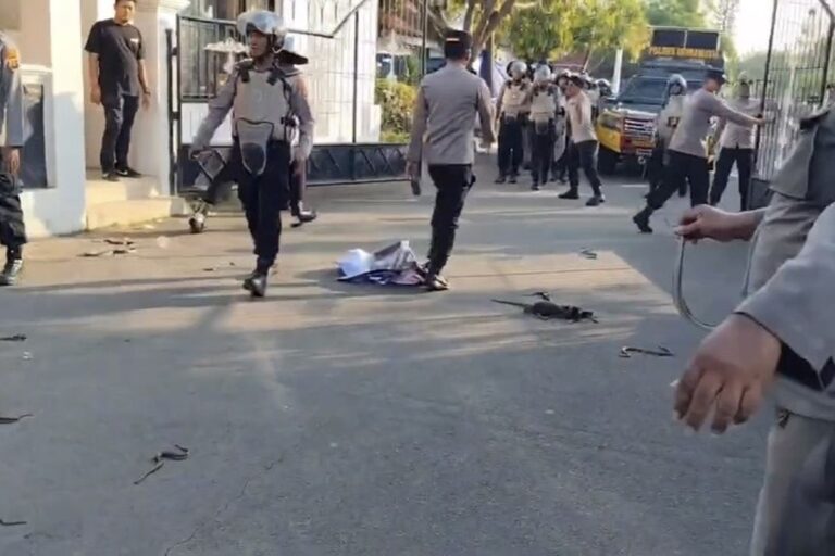Police officers in tactical gear on a street, debris scattered on the pavement, a crumpled cloth on the ground, and an officer in the foreground handling a rope or cord.