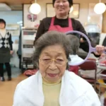 Elderly woman seated in a hair salon chair wearing a cape, smiling as staff prepare a round mirror for her styling.