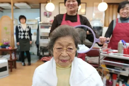 Elderly woman seated in a hair salon chair wearing a cape, smiling as staff prepare a round mirror for her styling.