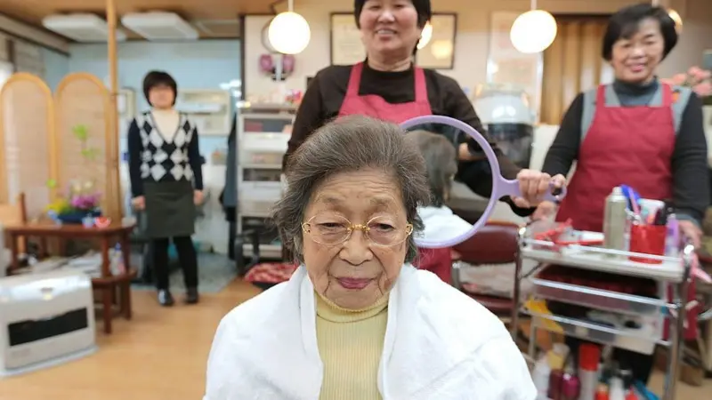 Elderly woman seated in a hair salon chair wearing a cape, smiling as staff prepare a round mirror for her styling.