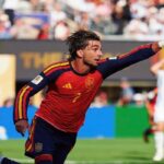 Soccer player in a red Spain kit celebrates by running and pointing toward the crowd on a sunny stadium field with fans in the background.