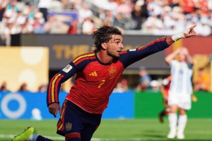 Soccer player in a red Spain kit celebrates by running and pointing toward the crowd on a sunny stadium field with fans in the background.