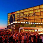 Crowd gathers outside the San Sebastián Film Festival venue at dusk, with the illuminated Donostia building and bold festival signage visible.