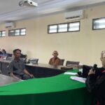 Panel discussion in a conference room: a woman in a hijab speaks into a microphone at a green-covered table, while attendees sit along the desks listening.