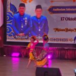 Children performing a colorful cultural dance on a stage with purple lights, wearing yellow skirts and pink or blue tops and flower headpieces; a large banner behind features officials in blue uniforms and the date 17 Oktober 2026.