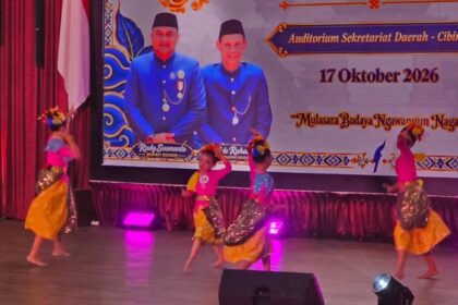 Children performing a colorful cultural dance on a stage with purple lights, wearing yellow skirts and pink or blue tops and flower headpieces; a large banner behind features officials in blue uniforms and the date 17 Oktober 2026.