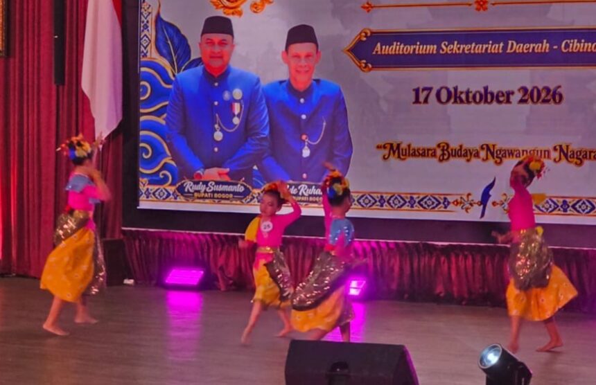 Children performing a colorful cultural dance on a stage with purple lights, wearing yellow skirts and pink or blue tops and flower headpieces; a large banner behind features officials in blue uniforms and the date 17 Oktober 2026.