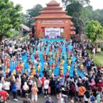 Crowd watches children in colorful traditional costumes perform a parade on a bright blue walkway in front of a brick stage at a cultural festival outdoors.