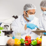 Two scientists in masks and hairnets examine peppers on a lab bench; one points at a clipboard held by the other, with a microscope nearby and plant shoots on the table.
