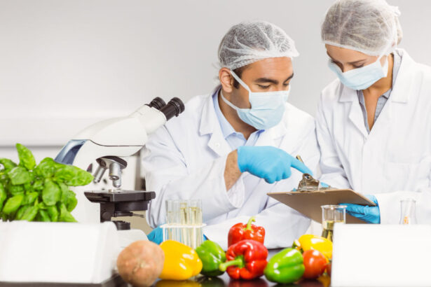 Two scientists in masks and hairnets examine peppers on a lab bench; one points at a clipboard held by the other, with a microscope nearby and plant shoots on the table.