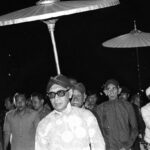 Man wearing sunglasses and a patterned kurta walks under ceremonial parasols at night, surrounded by a crowd of men.