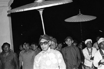 Man wearing sunglasses and a patterned kurta walks under ceremonial parasols at night, surrounded by a crowd of men.