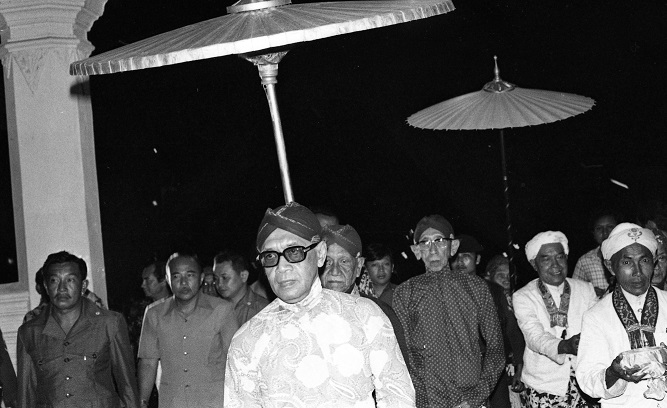 Man wearing sunglasses and a patterned kurta walks under ceremonial parasols at night, surrounded by a crowd of men.