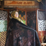 Woman in a hijab wearing a black embroidered shawl in a fabric shop, surrounded by colorful, patterned textiles on display.
