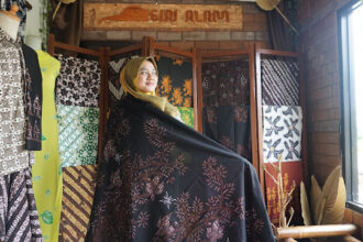 Woman in a hijab wearing a black embroidered shawl in a fabric shop, surrounded by colorful, patterned textiles on display.