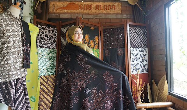 Woman in a hijab wearing a black embroidered shawl in a fabric shop, surrounded by colorful, patterned textiles on display.