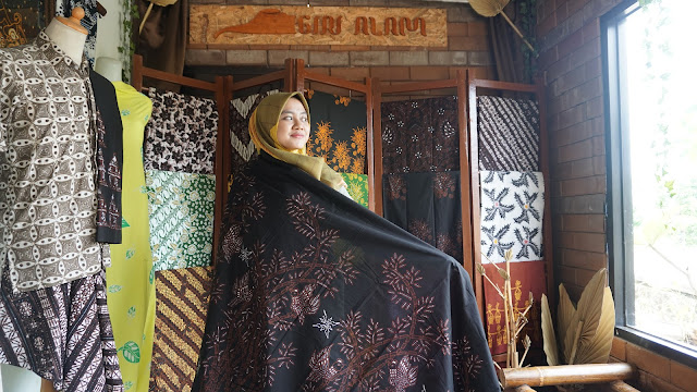 Woman in a hijab wearing a black embroidered shawl in a fabric shop, surrounded by colorful, patterned textiles on display.