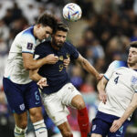 Three soccer players jump to head the ball during a match, with one in navy and two in white kits nearby.