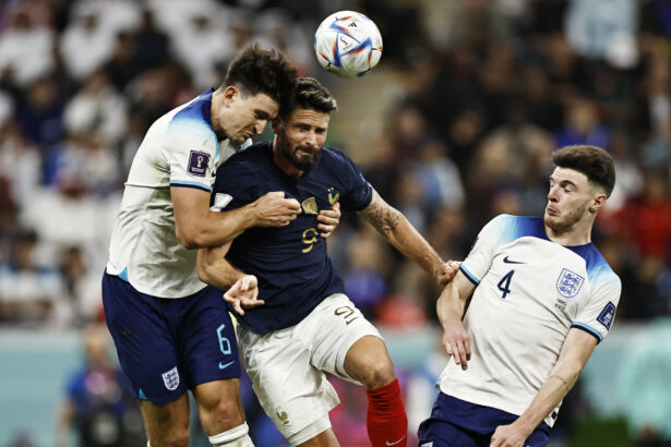 Three soccer players jump to head the ball during a match, with one in navy and two in white kits nearby.