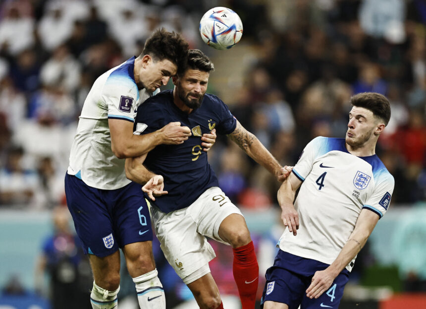 Three soccer players jump to head the ball during a match, with one in navy and two in white kits nearby.