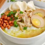Bowl of creamy congee topped with shredded chicken, halved soft-boiled eggs, peanuts, scallions, and crispy crackers.
