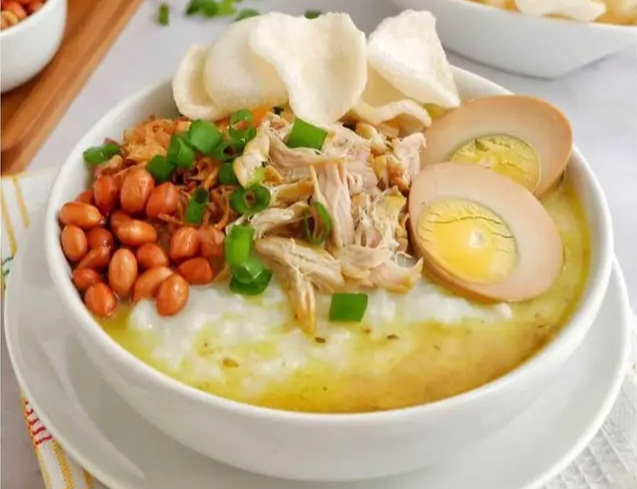 Bowl of creamy congee topped with shredded chicken, halved soft-boiled eggs, peanuts, scallions, and crispy crackers.
