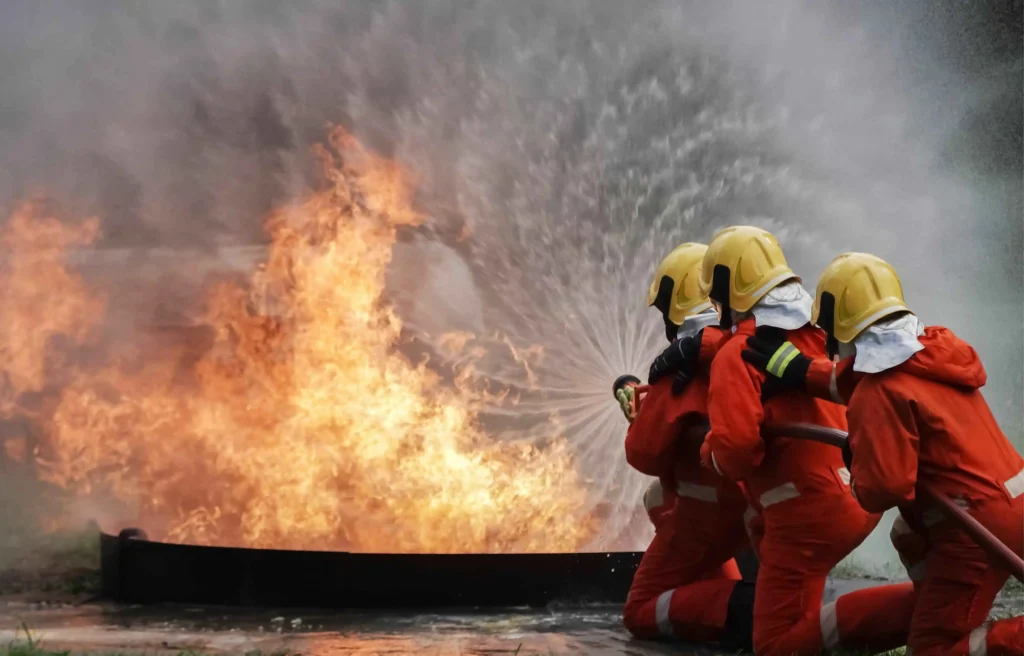 Firefighters in orange suits and yellow helmets aim a hose at large flames during a blaze.