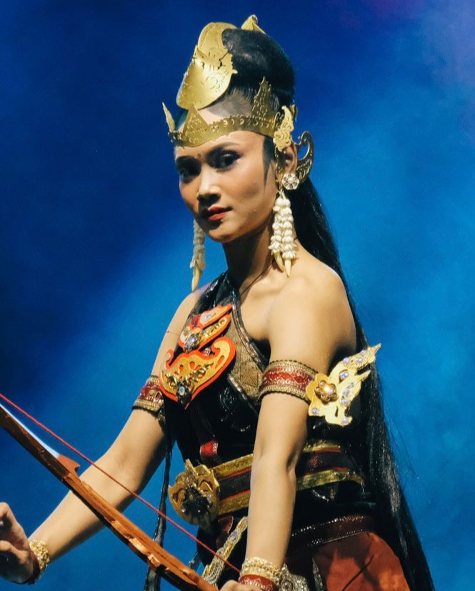 Female performer in an ornate gold headpiece and red/gold armor, holding a bow on a blue-lit stage background.