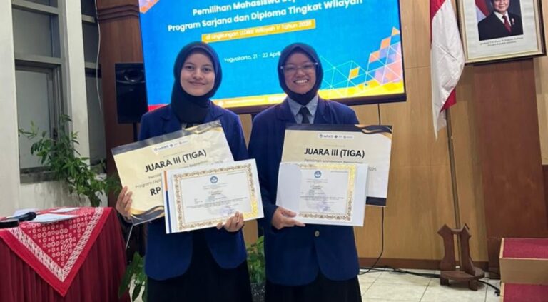 Two female students in blue blazers pose with certificates at a ceremony, smiling in front of a blue event screen and flags.