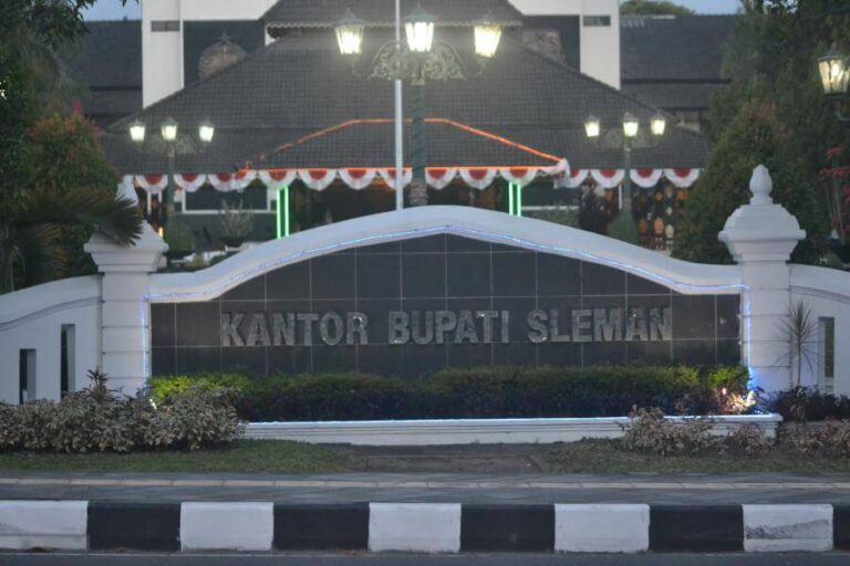 Signage at a gov’t building reading 'KANTOR BUPATI SLEMAN' behind a curved white wall with hedges and lights at dusk/uniformed garden area