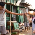 Girl in a blue shirt offering a small red fruit to a shirtless man on a dusty street market, during a sunny day.