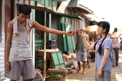 Girl in a blue shirt offering a small red fruit to a shirtless man on a dusty street market, during a sunny day.